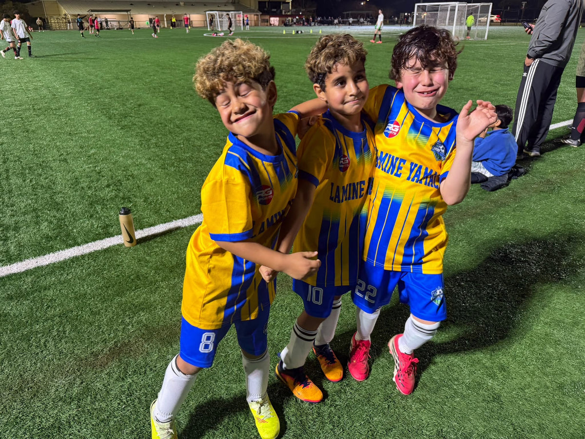 Three young soccer players in yellow and blue RG Sports team kits at a floodlit field